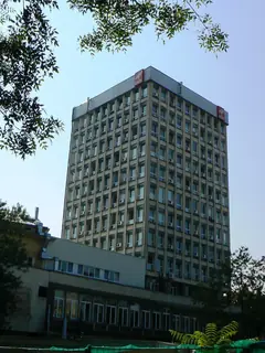 Висока многоетажна административна сграда с редици прозорци и климатици, с червени табели на покрива, сред клони на дървета и синьо небе