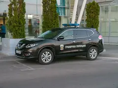 Тъмен автомобил на Гранична полиция със синя сигнална лампа, паркиран пред стъклена сграда с туи